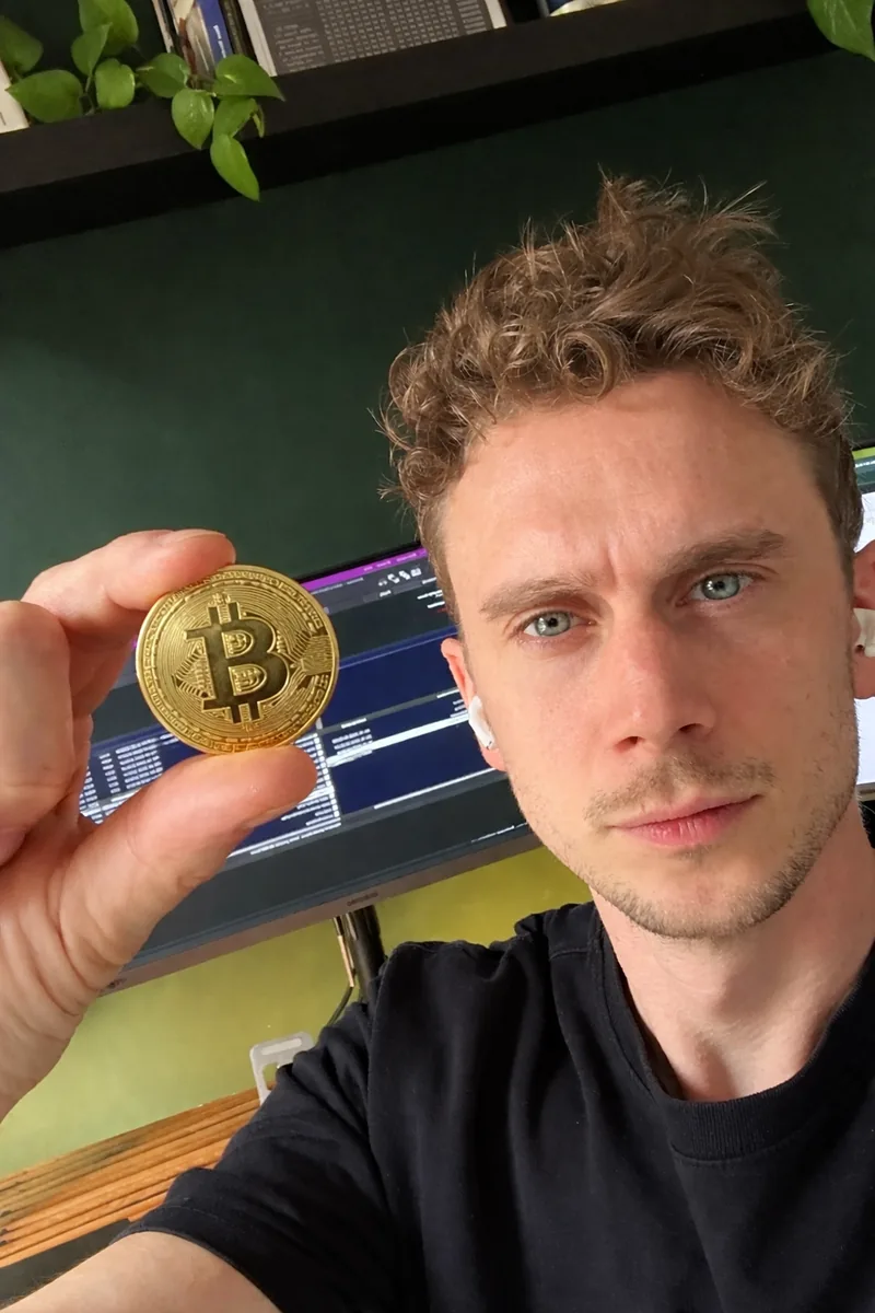 Paul Brock houdt een fysieke bitcoin vast, met handelsschermen op de achtergrond